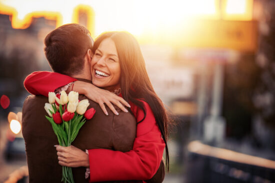 A man and woman embrace while holding flowers and smiling.