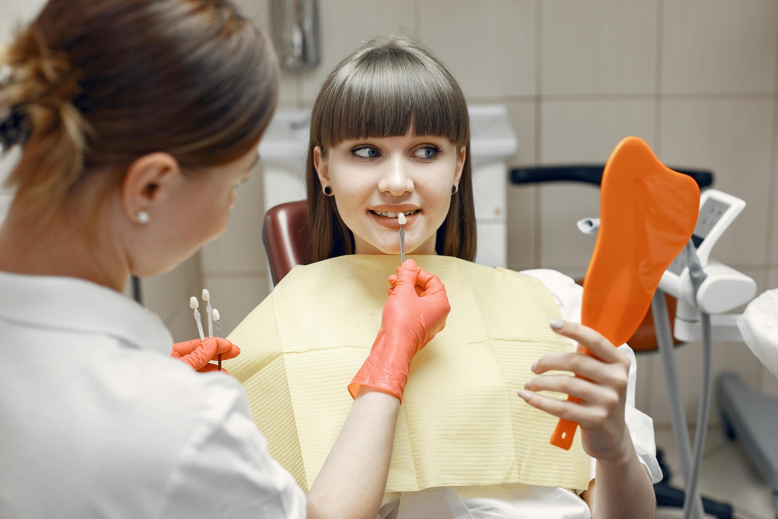 A woman sits in a dentist chair, holding a dental tool, preparing for a veneers procedure.