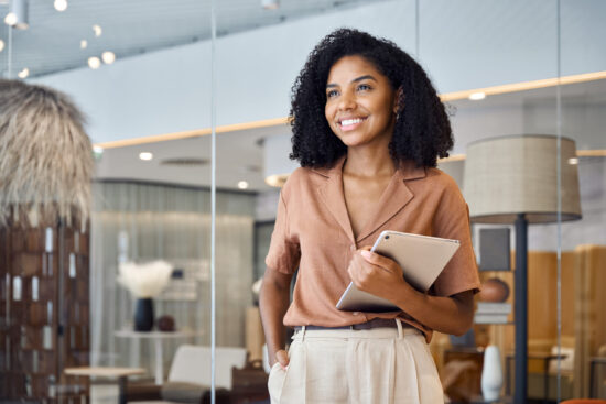 A woman smiles while holding a tablet in an office setting.